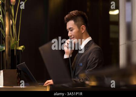 Ein lächelnder chinesischer männlicher Rezeptionist spricht am Telefon an der Hotelrezeption. Die Szene vermittelt professionelle Gastfreundschaft und effizienten Service, ideal für Reisen und geschäftliche Umgebungen. Stockfoto