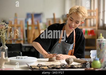 Ein glücklicher junger asiatischer Mann bastelt Keramikkeramik und kntet Ton in einer Handwerkstatt. Die Szene zeigt Kreativität und handwerkliche Fähigkeiten, die für Kunstkurse, Studios und inklusives Hobby geeignet sind. Stockfoto