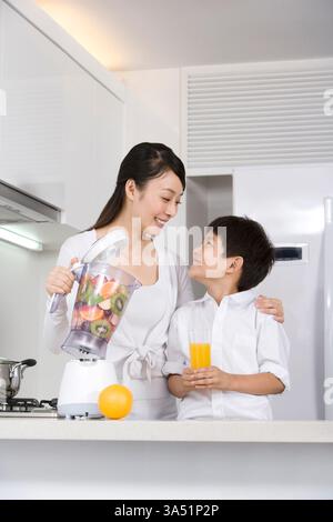 Die chinesische Mutter und ihr Sohn bereiten frischen Orangensaft in der Küche zu und verwenden einen Mixer für ein gesundes Getränk. Die Szene betont gesunde Familienroutinen und das häusliche Leben. Ideal für Lifestyle, Ernährung und Hausmannskost. Stockfoto
