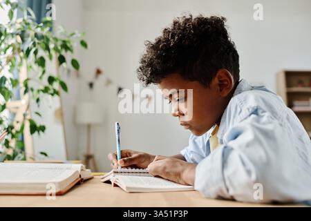Afrikanischer Grundschüler, der Hausaufgaben zu Hause macht, an einem Tisch mit Buch und Notizbuch sitzt. Dieser konzentrierte Lernmoment spiegelt Bildung und Unterstützung durch die Familie wider. Stockfoto