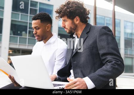Konzentrierter hispanischer Geschäftsmann mit Dokumenten, der mit einem männlichen Kollegen aus kaukasien arbeitet, während er auf einer Bank vor dem Bürogebäude sitzt Stockfoto