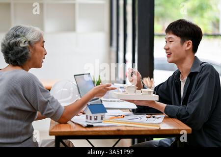 Ältere asiatische Frau, die sich mit einem asiatischen Ingenieur trifft, der das Modellhaus hält und bespricht, während sie am Tisch im Büro sitzt Stockfoto