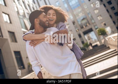 Fröhliche Afroamerikanerin mit lockigen Haaren steht auf einer sonnigen Stadtstraße und umschließt ihren Freund von hinten. Perfekt für Romantik, Beziehung und urbanen Lifestyle. Gut beleuchtete Straßenszene im Freien. Stockfoto