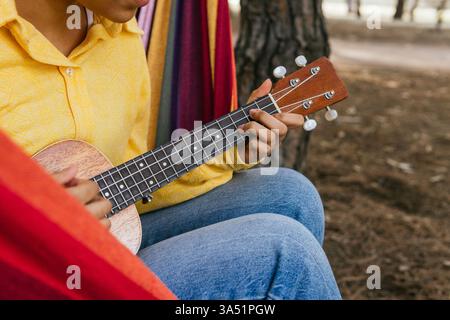 Abgeschnittene, nicht erkennbare Frau in einem hellen Hemd, die in einer Park-Hängematte liegt, während sie Ukulele spielt. Dieser unbeschwerte Outdoor-Moment passt zu Lifestyle, Musik und Freizeitkampagnen. Stockfoto