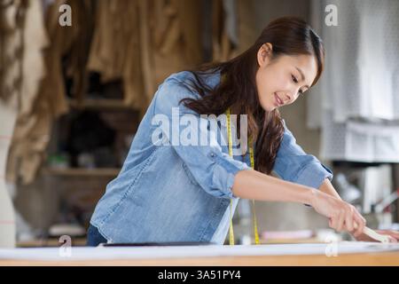 Chinesischer Modedesigner arbeitet in einem Designstudio. Umgeben von Stoffen und Werkzeugen konzentriert sie sich auf den kreativen Prozess. Stockfoto