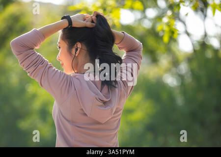 Eine Chinesin bindet sich die Haare, um sich auf einen morgendlichen Lauf im Park vorzubereiten. Energiegeladenes Lifestyle-Image mit Fitness, Outdoor-Training und tragbaren Technologien wie Ohrstöpseln und Smartwatch. Perfekt für gesundes Leben, Wellness und urbane Laufkampagnen. Stockfoto