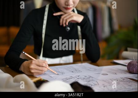 Nahaufnahme einer professionellen asiatischen Modedesignerin, die in ihrem Studio eine neue Kollektion zeichnet und entwirft. Dieses fokussierte Porträt hebt Kreativität, Modedesign und Fachwissen hervor. Ideal für Boutique-Studios, Designkarrieren und Mode für kleine Unternehmen. Stockfoto