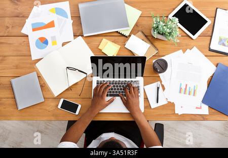 Blick von oben auf einen afroamerikanischen Geschäftsmann an einem Holzschreibtisch, der in einem Büro an einem Laptop arbeitet. Die Zusammensetzung der Draufsicht hebt einen ausgelasteten Arbeitsbereich mit Kopierbereich für Messaging hervor. Geeignet für Produktivität, Technologie und Unternehmenskommunikation. Stockfoto