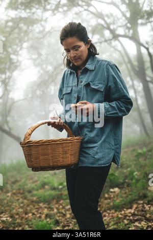Latina-Frau im Wald sammelt wilde Pilze, trägt einen Korb in trüben Waldgebieten. Eine natürliche Outdoor-Szene, ideal für Natur, Nahrungssuche und ländliche Lifestyle-Themen. Ideal für Artikel über saisonale Ernte und nachhaltige Lebensmittelbeschaffung. Stockfoto