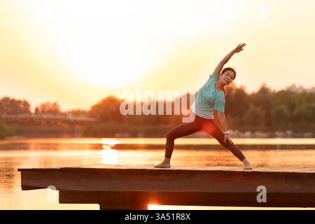 Chinesische Frau mittleren Alters führt Yoga auf einem hölzernen Pier bei Sonnenuntergang durch, im Freien vom Wasser gefangen. Die hinterleuchtete Szene betont Ruhe, Balance und Entspannung in einer ruhigen Umgebung am Fluss. Ideal für Wellness-, Fitness- und Lifestyle-Kampagnen mit Achtsamkeit, Outdoor-Übungen und asiatischen Themen. Stockfoto