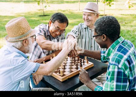 Ein lächelnder älterer asiatischer Mann sitzt mit einer Gruppe älterer Männer an einem sonnigen Tag in einem Park. Sie versammeln sich um einen Tisch, genießen Outdoor-Freizeit und Freundschaft. Stockfoto