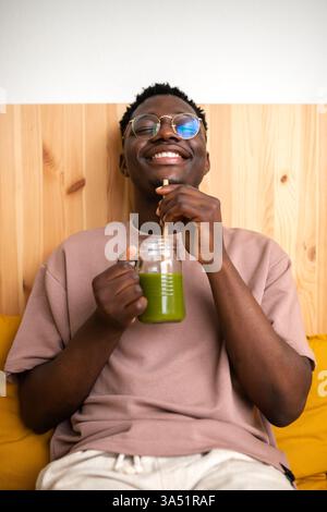 Entspannter Lifestyle Aufnahme eines Schwarzen Mannes, der zu Hause einen gesunden grünen Saft genießt. Er schlürft von einem Bambusstroh und vermittelt Wohlbefinden und achtsames Leben. Geeignet für Artikel über gesunde Gewohnheiten, Entgiftung und Wellness-Kampagnen zu Hause. Stockfoto