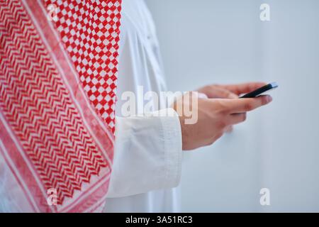 Porträt eines jungen arabischen muslimischen Geschäftsmannes, der ein Smartphone in einem modernen technischen Kontext benutzt. Ideal für Unternehmen, Unternehmergeist und digitale Kommunikationsgrafiken. Geeignet für Märkte im Nahen Osten, traditionelle Kleidung und das Berufsleben. Stockfoto