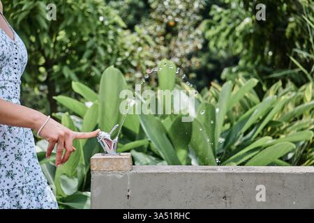 Eine junge Frau im Sommerkleid trinkt während eines Spaziergangs Wasser aus einem Springbrunnen im Park. Outdoor-Lifestyle-Bild für Erfrischung und Freizeit an einem sonnigen Tag. Ideal für Wellness-, Sommer- und Freizeitkampagnen im Freien. Stockfoto