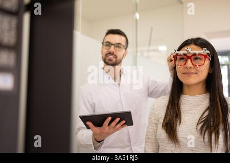 In einem optischen Geschäft versucht eine hispanische Frau eine Brille an, während ein kaukasischer Optiker einen Versuchsrahmen anpasst. Die Szene konzentriert sich auf Sehprüfungen, die Rahmenauswahl und die Patientenbetreuung in einem vielfältigen Brillengeschäft. Ideal für Optometrie, Brilleneinkäufe und Einzelhandelsbilder. Stockfoto