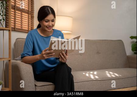 Eine Asiatin sitzt bequem auf einem Sofa in einem gemütlichen Wohnzimmer und liest ein Buch mit einem Lächeln. Diese warme Wohnszene beleuchtet Freizeit, Lernen und den Alltag zu Hause in einem modernen Apartment. Stockfoto
