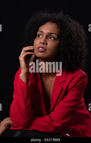 Studio-Porträt einer nachdenklichen jungen schwarzen Frau mit einem Afro im roten Mantel. Sie sitzt vor schwarzem Hintergrund und blickt weg, um eine nachdenkliche Stimmung zu erzeugen. Ideal für Mode-, Beauty- und Lifestyle-Projekte mit selbstbewussten, unabhängigen Schwarzen Frauen. Stockfoto