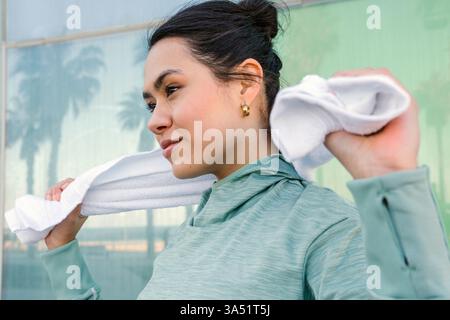 Die lächelnde hispanische Frau in Fitnesskleidung hält draußen ein Handtuch neben einer Glaswand. Die Szene vermittelt Energie, Gesundheit und aktives Leben für Wellness-Kampagnen. Stockfoto