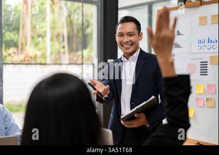 Eine selbstbewusste asiatische Führungskraft leitet ein Meeting in einem Sitzungssaal und bittet Kollegen um Fragen. Diese professionelle Szene eignet sich ideal für Unternehmenskommunikation, Teamarbeit und Führungskampagnen. Stockfoto