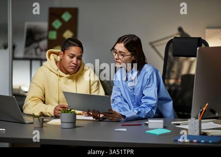Zwei junge Frauen diskutieren nachts in einem modernen Büro über Dokumente. Die Szene konzentriert sich auf Teamarbeit, Überstunden und einen professionellen, vielfältigen Arbeitsplatz. Ideal für Unternehmen, Frauen am Arbeitsplatz und Collaboration-Bilder. Stockfoto