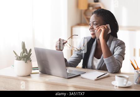 Selbstbewusste Afro-Geschäftsfrau auf einem Handy im modernen Büro, mit stilvoller Brille und Kopierraum. Ein eindrucksvolles Porträt weiblicher Führungs- und Unternehmertätigkeit für Unternehmens-, Marketing- oder Personalkampagnen. Ideal für Schwarzweißbilder und professionelle Lifestyle-Bilder. Stockfoto