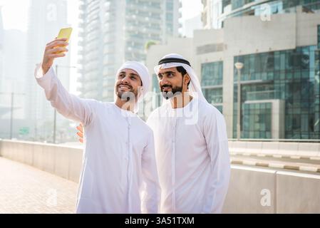 Lächelnde Männer aus dem Nahen Osten in Kanduras machen ein Selfie auf einer sonnigen Stadtstraße. Ein zeitgenössischer urbaner Moment, ideal für geschäftliche Netzwerke und Kampagnen zur modernen arabischen Kultur. Stockfoto