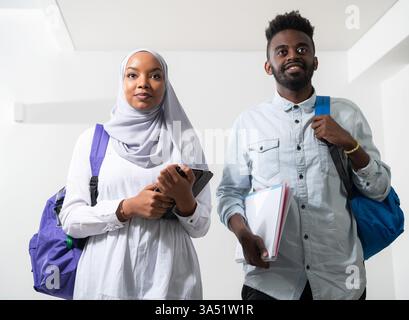 Junge afrikanische Studenten, darunter eine Frau mit traditionellem Hijab, gehen als ein vielfältiges Geschäftsteam zusammen, isoliert auf weißem Hintergrund. Das Image betont Vielfalt, Bildung und Teamarbeit. Stockfoto