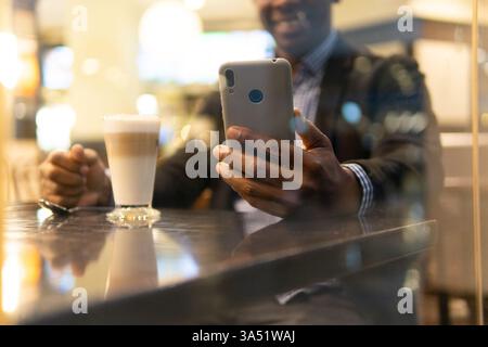 Ein schwarzer Geschäftsmann in einem Café benutzt ein Smartphone und ein Glas Getränk in der Nähe. Die Szene fängt einen Moment des Geschäfts und der Konnektivität in einem entspannten, modernen Ambiente ein. Stockfoto