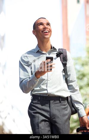 Porträt eines glücklichen jungen Mannes, der mit Handy und Tasche durch die Stadt geht. Urbanes Lifestyle-Bild, das Selbstvertrauen, Konnektivität und Alltagsstil vermittelt. Stockfoto