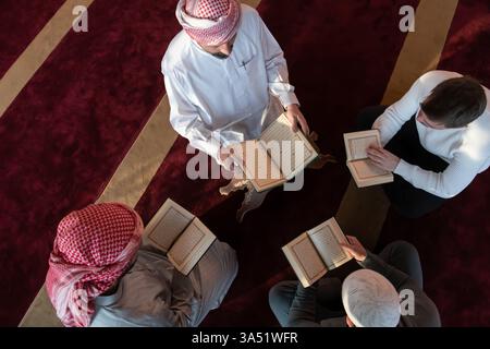 Blick von oben auf eine vielfältige Gruppe von Muslimen, die gemeinsam den Koran in einer Moschee lesen. Diese islamische Bildungsszene betont Hingabe, Studium und spirituelle Praxis. Stockfoto