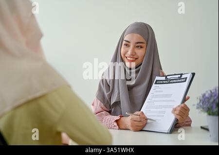 Eine junge muslimische Immobilienmaklerin erklärt ihrem Kunden in einem professionellen Treffen die Vereinbarung für einen Immobilienkauf. Die Szene zeigt Immobiliendienstleistungen, Vertragsunterzeichnung und Kundenberatung. Ideal für die Vermarktung von Immobilien, Immobilienmärkten und Immobilienagenturen mit asiatischen Fachleuten. Stockfoto