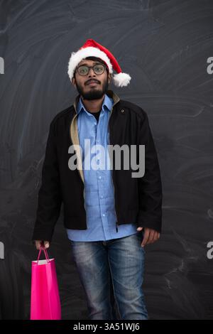 Ein indischer Mann in festlicher Partykleidung trägt einen Weihnachtsmann-Hut und hält Einkaufstaschen vor einem dunklen Studio-Hintergrund. Dieses fröhliche Weihnachtsbild zeigt Weihnachtseinkäufe, Geschenke und fröhliche Feierlichkeiten. Stockfoto