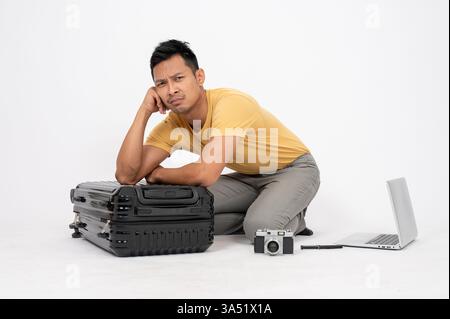 Trauriger asiatischer Mann, der sich auf dem Boden vor weißem Hintergrund im Studio auf das Gepäck gestützt hat Stockfoto