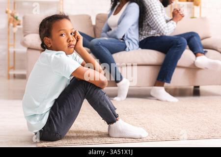 Porträt eines traurigen afroamerikanischen Mädchens, das nach einem Streit zu Hause neben ihren Eltern sitzt. Dieses ergreifende Familienkonzept vermittelt Konflikte, Einsamkeit und die Auswirkungen von häuslichem Stress. Geeignet für Artikel oder Kampagnen zu Familie, Elternschaft, Beziehungen oder geistigem Wohlbefinden. Stockfoto