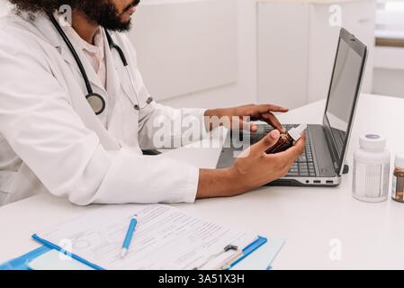 Ein ernsthafter bärtiger männlicher Arzt sitzt an einem Krankenhaustisch mit einem Stethoskop und einer Flasche Medizin zur Hand. Dieses professionelle Gesundheitsbild vermittelt medizinische Beratung und Patientenversorgung in einem klinischen Umfeld. Stockfoto