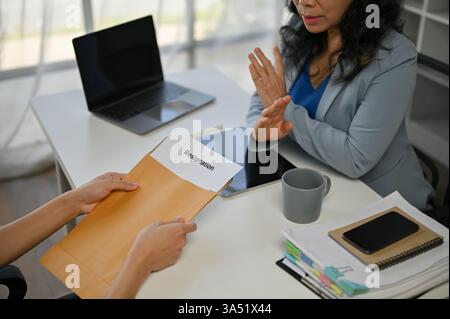 Asiatischer männlicher Arbeiter, der ernsthafte Gespräche führt und asiatische Geschäftsfrau einen Rücktrittsbrief übergibt, während er im Büro auf einem Tisch sitzt Stockfoto