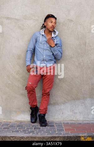 Ganzkörperporträt eines selbstbewussten afrikanischen Mannes, der sich in einer städtischen Umgebung an eine Wand lehnt. Dieser stilvolle, legere Look betont Street-Style und maskuline Mode und ist ideal für Lifestyle- oder Modebilder. Stockfoto