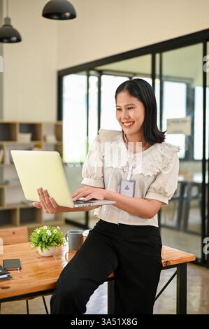 Porträt einer selbstbewussten thailändischen Geschäftsfrau in einem modernen Büro, die einen Laptop hält. Ihre ausgefeilte Haltung vermittelt Professionalität und Führung in einem Unternehmensumfeld. Ideal für Geschäfts-, Arbeitsplatz- und Technologiethemen mit Frauen im Management. Stockfoto