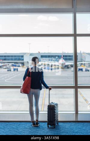 Frau mit Tasche, die auf das Fenster im Flughafen blickt, während sie ihr Gepäck hält Stockfoto