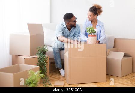 Glückliches afroamerikanisches Paar sitzt drinnen auf einer Couch und packt Umzugskartons für den Umzug. Dank der selektiven Fokussierung und des Kopierbereichs ist es vielseitig für Heim- und Immobilienmotive geeignet. Ideal für Umzüge, Standortwechsel und Bilder für den Familienleben. Stockfoto