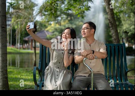 Eine hübsche und glückliche asiatische Frau und ihr Vater machen ein Selfie auf einer Bank in einem grünen Park. Sie lächeln zusammen und halten einen fröhlichen Familienmoment auf einem Smartphone fest. Perfekt für Familien-, Zweisamkeit- oder Lifestyle-Kampagnen. Stockfoto