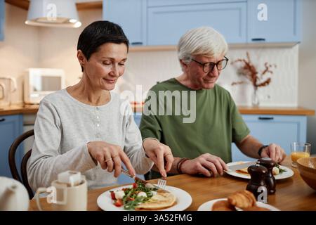 Seniorenpaar genießen das gemeinsame Frühstück zu Hause, beide sehen zufrieden aus, während sie am Holztisch in komfortabler Küche essen und gegenseitigen Glück und Verbundenheit zeigen Stockfoto