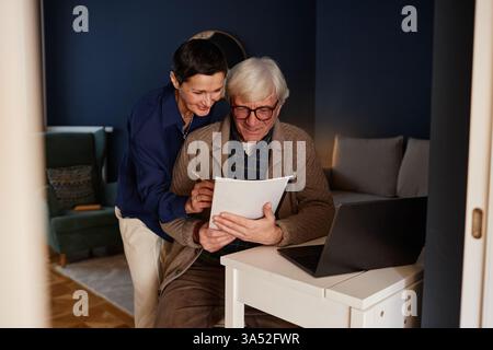 Lächelnde Frau, die einem älteren Mann beim Schreiben auf Papier vor dem Laptop auf dem Schreibtisch hilft. Gemütliches Zimmer mit komfortablen Sitzgelegenheiten und warmer Beleuchtung für eine angenehme Atmosphäre Stockfoto