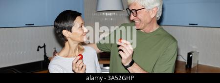 Die Kopfzeile der Website zeigt, wie ein lächelndes Paar einen glücklichen Moment teilt, während er Kirschtomaten hält, die in der Küche kochen Stockfoto