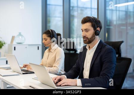 Zwei Kundendienstmitarbeiter, ein Mann und eine Frau, die Headsets tragen, unterstützen Kunden bei der Nutzung ihres Laptops in einem gut beleuchteten, modernen Büro. Stockfoto