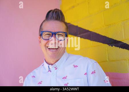 Ein lächelnder Mann mit kurzen, grauen Haaren, blauen Brillen und einem hellblauen Hemd mit Flamingomuster steht vor einem rosa und gelben Ziegelstein Stockfoto