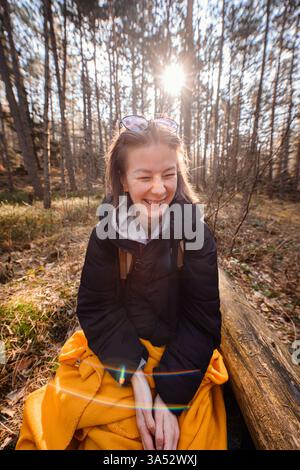 Die lächelnde junge Frau in einer schwarzen Jacke sitzt auf einem Baumstamm in einem sonnendurchfluteten Wald, umhüllt in eine gelbe Decke, genießt Natur und Wärme. Stockfoto