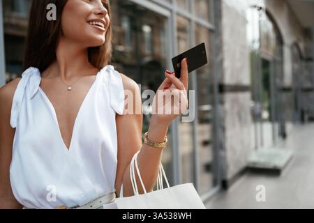 Kreditkarte liegt in der Hand. Stilvolle Frau mit Einkaufstasche draußen in der Nähe des Gebäudes. Stockfoto