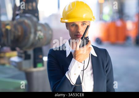 Frau mit Schutzhelm, die Walkie-Talkie benutzt und die Geschäfte im Lager verwaltet Stockfoto