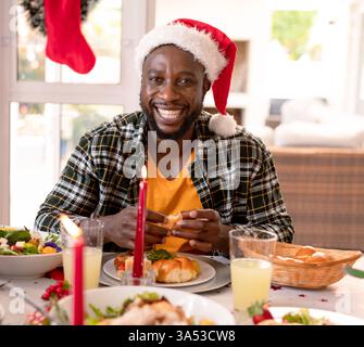 Ein lächelnder Mann mit Weihnachtsmann-Hut genießt festliches Essen zu Hause mit der Familie Stockfoto